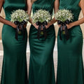 Three bridesmaids in emerald green satin dresses holding white baby's breath bouquets with black ribbon bows