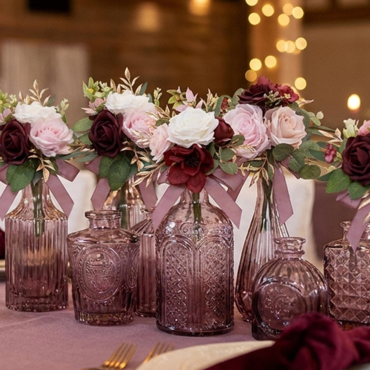 Close-up of mauve pressed-glass bud vases with pink, white and burgundy roses tied with dusty rose ribbon